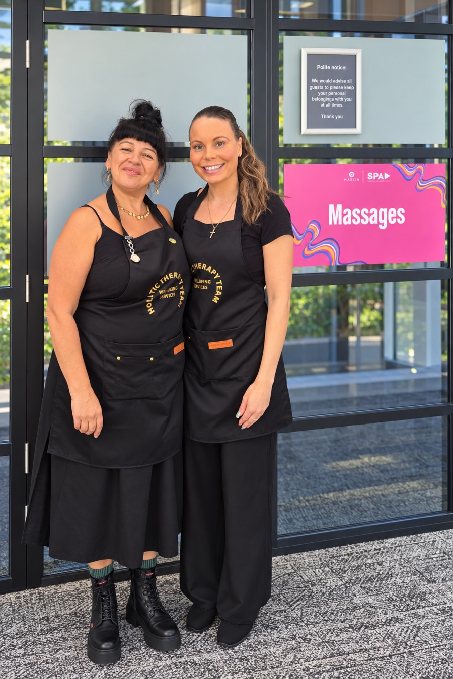 Two KM therapists in black Holistic Therapy Team aprons beside a pink Massages sign at a client's offices