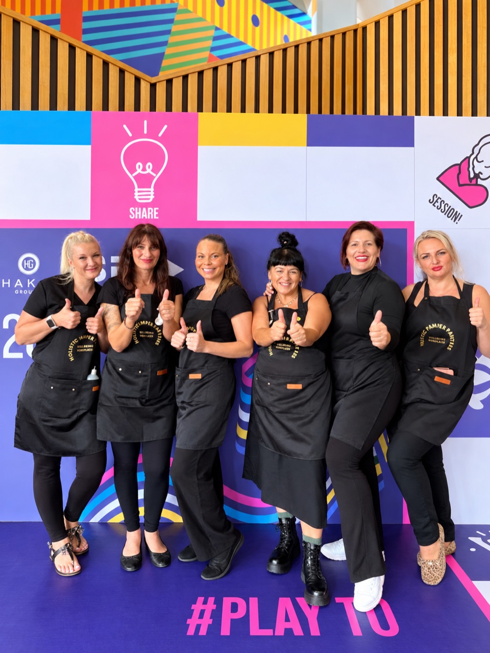 Six KM therapists in black aprons giving a thumbs up in front of the colourful Hakim SPA backdrop